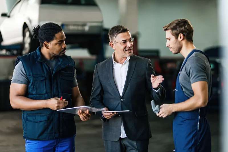 A man in a suit talking with two mechanics in a repair shop bay