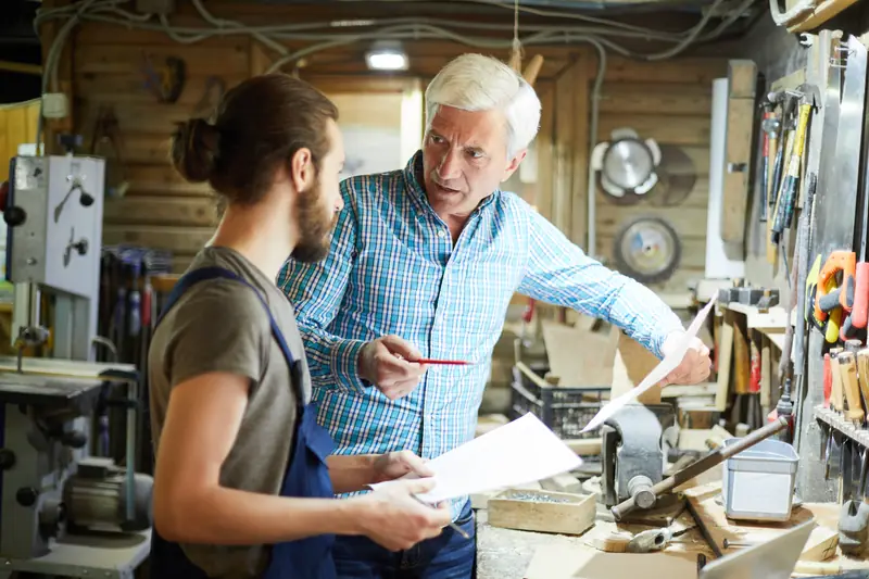 An older man going over a sheet of notes with a younger worker in a workshop