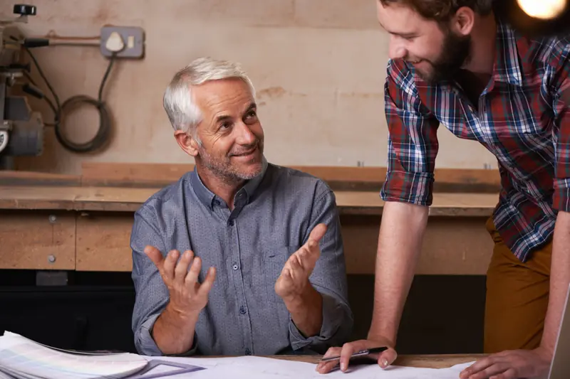 A seated man talking through papers on a workbench with a colleague leaning in beside him