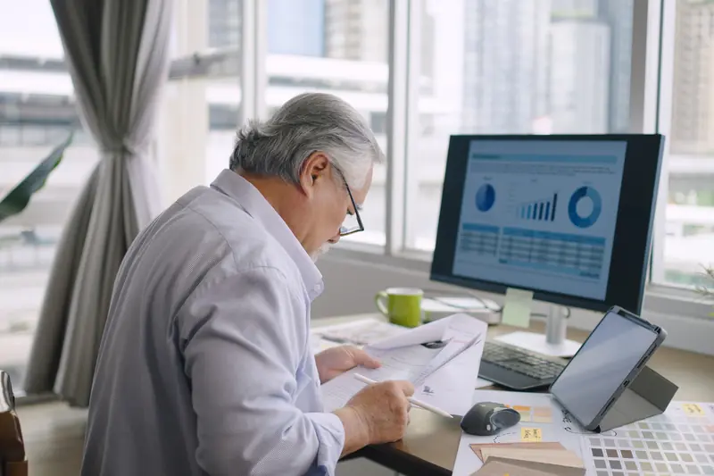 A man at his desk reading printed pages beside a monitor full of charts