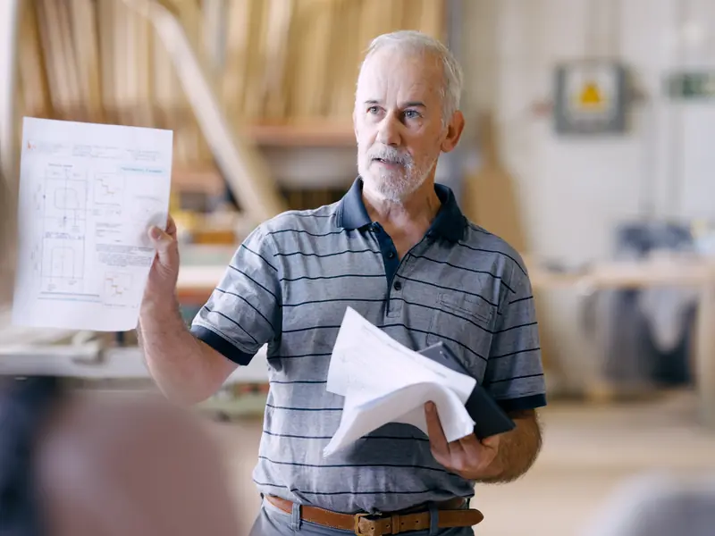 A man holding up a printed plan while he talks to a small group in a workshop